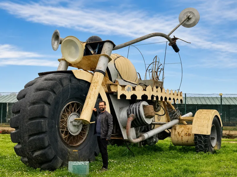 A man stands next to a large, artistic vehicle with oversized tires and a unique design, set against a blue sky.