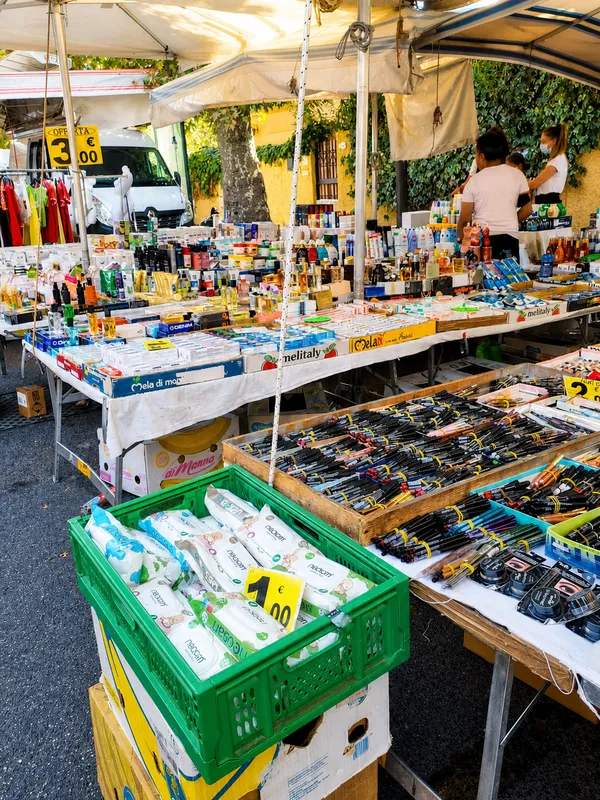 A bustling market stall displays various products, including cosmetics, pens, and household items, under a canopy.